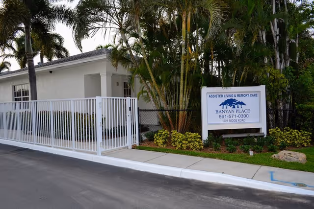 Exterior view of Banyan Place assisted living and memory care facility showing a white building partially obscured by palm trees and greenery, a white metal fence, and a sign with the facility's name, phone number, and address.
