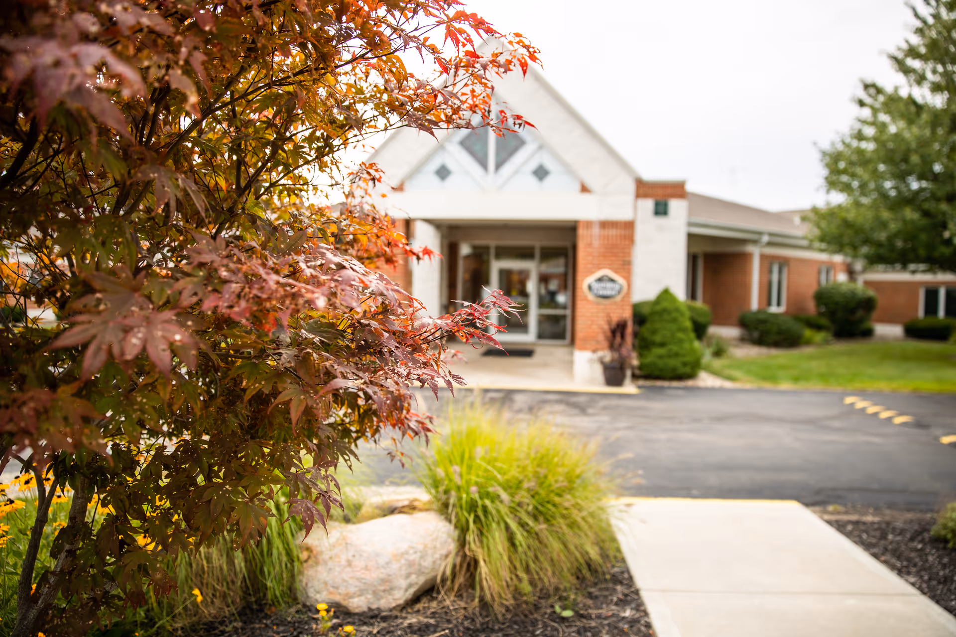 View of the entrance to a brick building with a triangular roof section above the doorway, surrounded by landscaped greenery including a tree with red leaves and ornamental grasses.
