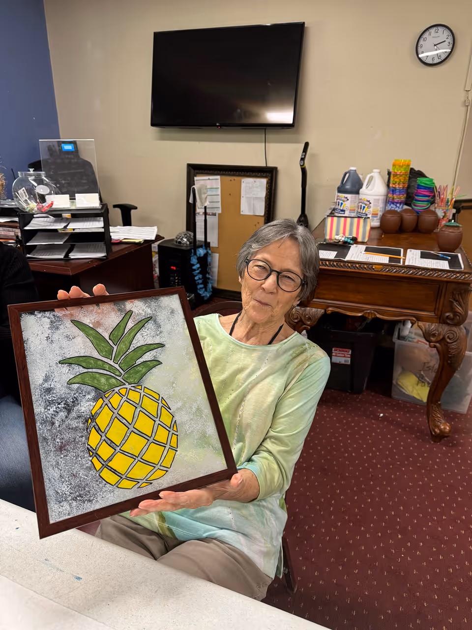 An elderly woman with short gray hair and glasses is sitting indoors, holding up a framed artwork of a yellow pineapple with green leaves. Behind her is a wall-mounted TV, a clock, and a wooden table with various items including bottles, colorful cups, and papers.