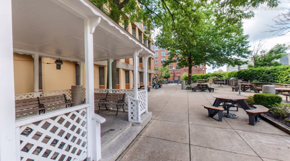 Outdoor courtyard area with picnic tables and benches, a covered seating area with wooden benches, trees, and surrounding buildings under a partly cloudy sky.