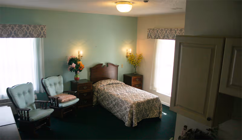 A cozy bedroom with a single bed covered in a patterned bedspread, two cushioned wooden chairs, two nightstands with floral arrangements, wall-mounted lamps, and two windows with sheer curtains and valances.
