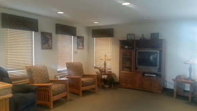 A cozy living room with upholstered armchairs, wooden side tables and a TV cabinet beneath three windows with blinds.