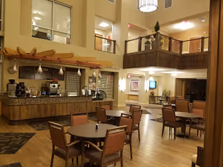 Interior view of a spacious common area in a senior living facility featuring a coffee and snack bar with a granite countertop, several round tables with chairs, and a second-floor balcony overlooking the area. The space is well-lit with ceiling lights and decorated with plants and framed artwork.