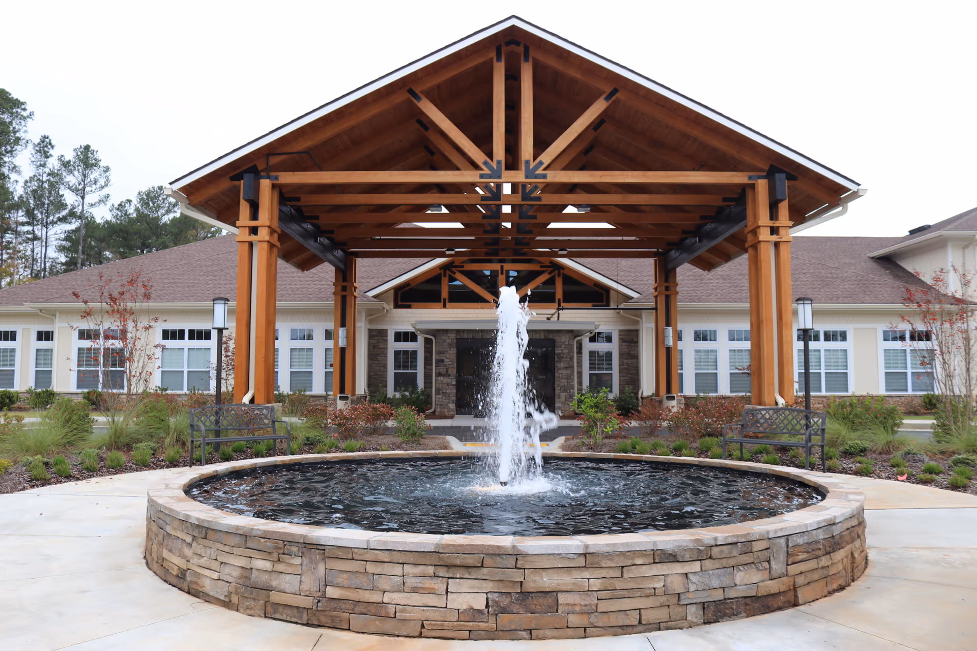 Outdoor view of Oaks at Grove Park featuring a circular stone fountain with water spraying upwards, two metal benches on either side, and a large wooden pergola structure in front of a building with multiple windows and a sloped roof.