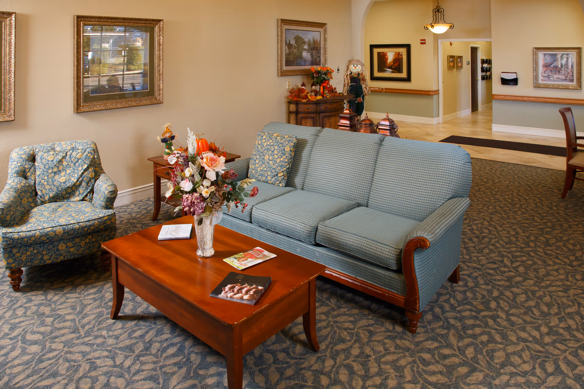 A cozy living room area with a blue patterned sofa and matching armchair. A wooden coffee table with a floral arrangement and magazines sits in front of the sofa. The room has framed artwork on the walls, a patterned carpet, and a small wooden side table. In the background, there is a hallway and a wooden cabinet decorated with autumn-themed items and a scarecrow figure.