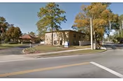 Corner view of a single-story beige building surrounded by trees and lawns at the edge of a street.