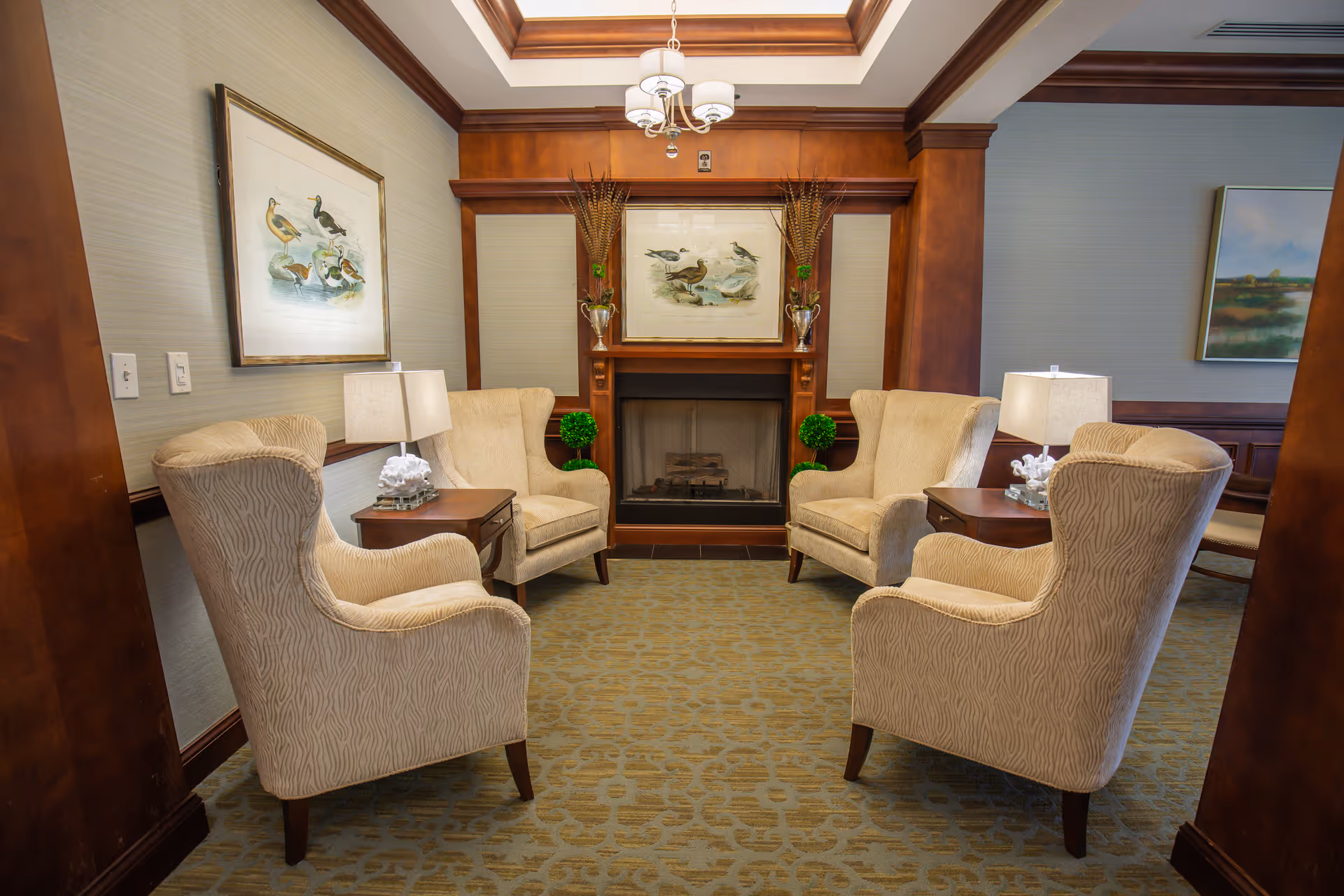 A cozy seating area in a senior living facility with four beige upholstered armchairs arranged around two wooden side tables with lamps. The room features a wooden fireplace with framed bird artwork above it, decorative plants, and a patterned carpet. The walls are light-colored with wood trim, and there is a skylight above providing natural light.