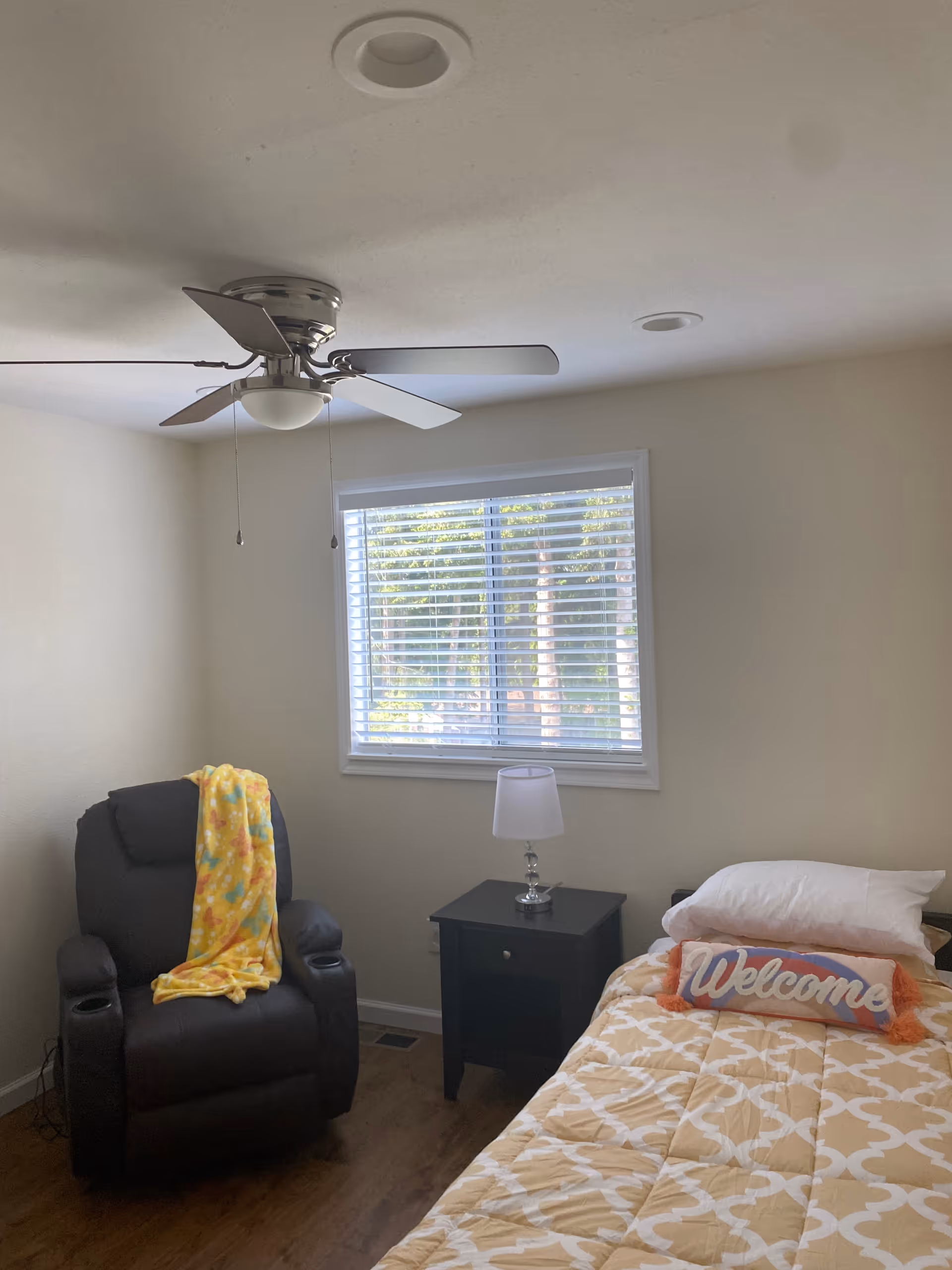 A cozy bedroom with a single bed covered in a yellow patterned comforter and a pillow with a 'Welcome' sign. Next to the bed is a black nightstand with a small lamp. A black recliner chair with a yellow blanket draped over it is positioned near a window with white blinds. The ceiling has a silver ceiling fan with five blades.