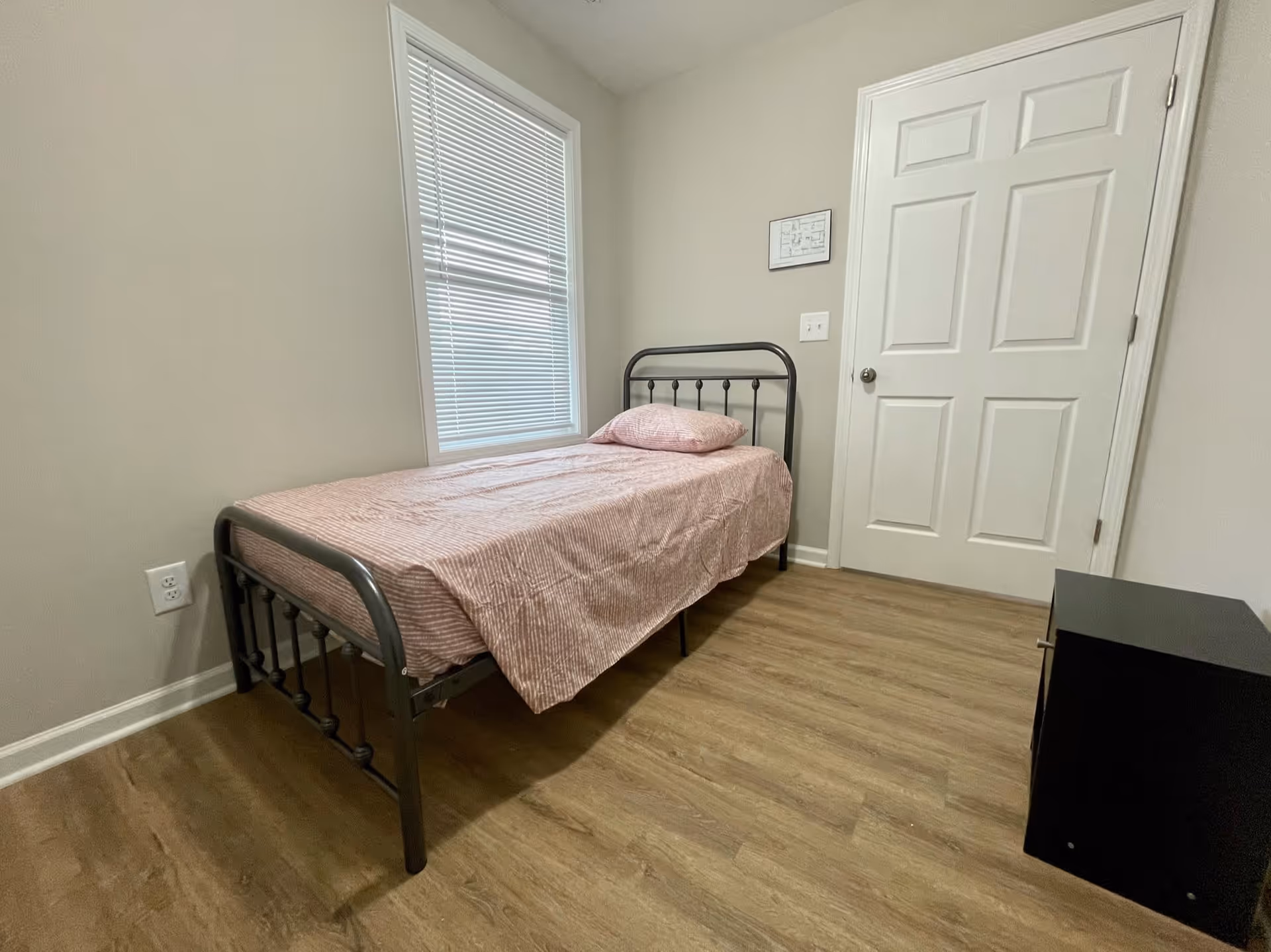 A small bedroom with a single metal bed frame, a pink striped bedspread and pillow, a window with closed blinds, a white door, and a small black nightstand on a wooden floor.