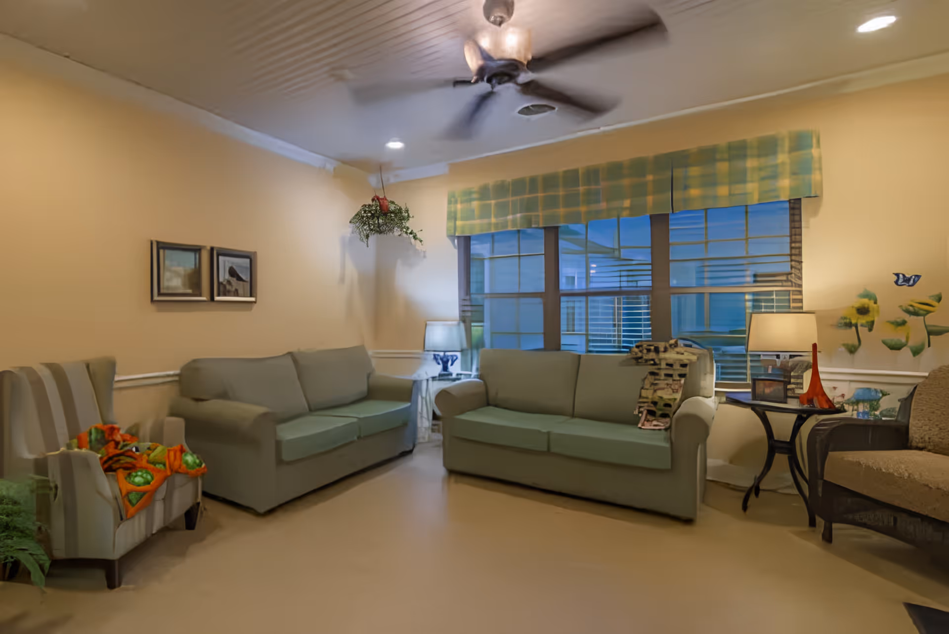 Communal living room with two green sofas, armchairs, side tables and lamps in front of a large window and ceiling fan.