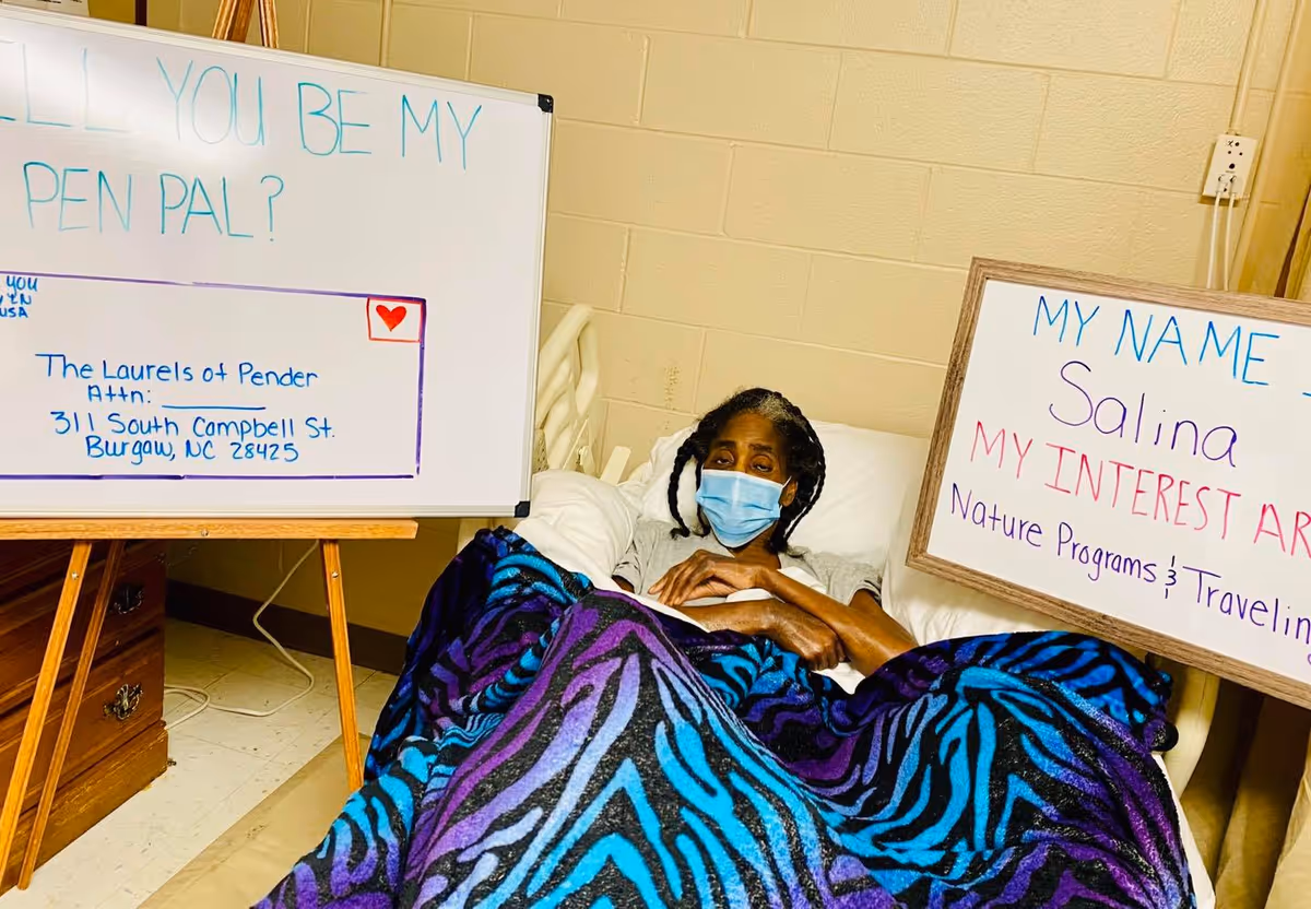 An elderly woman wearing a face mask is lying in a bed covered with a blue and purple patterned blanket. She is holding a whiteboard that says 'MY NAME Salina MY INTEREST ARE Nature Programs & Traveling'. Next to her is another whiteboard on an easel that reads 'WILL YOU BE MY PEN PAL? The Laurels of Pender Attn: 311 South Campbell St. Burgaw, NC 28425'. The room has beige walls and a wooden nightstand.