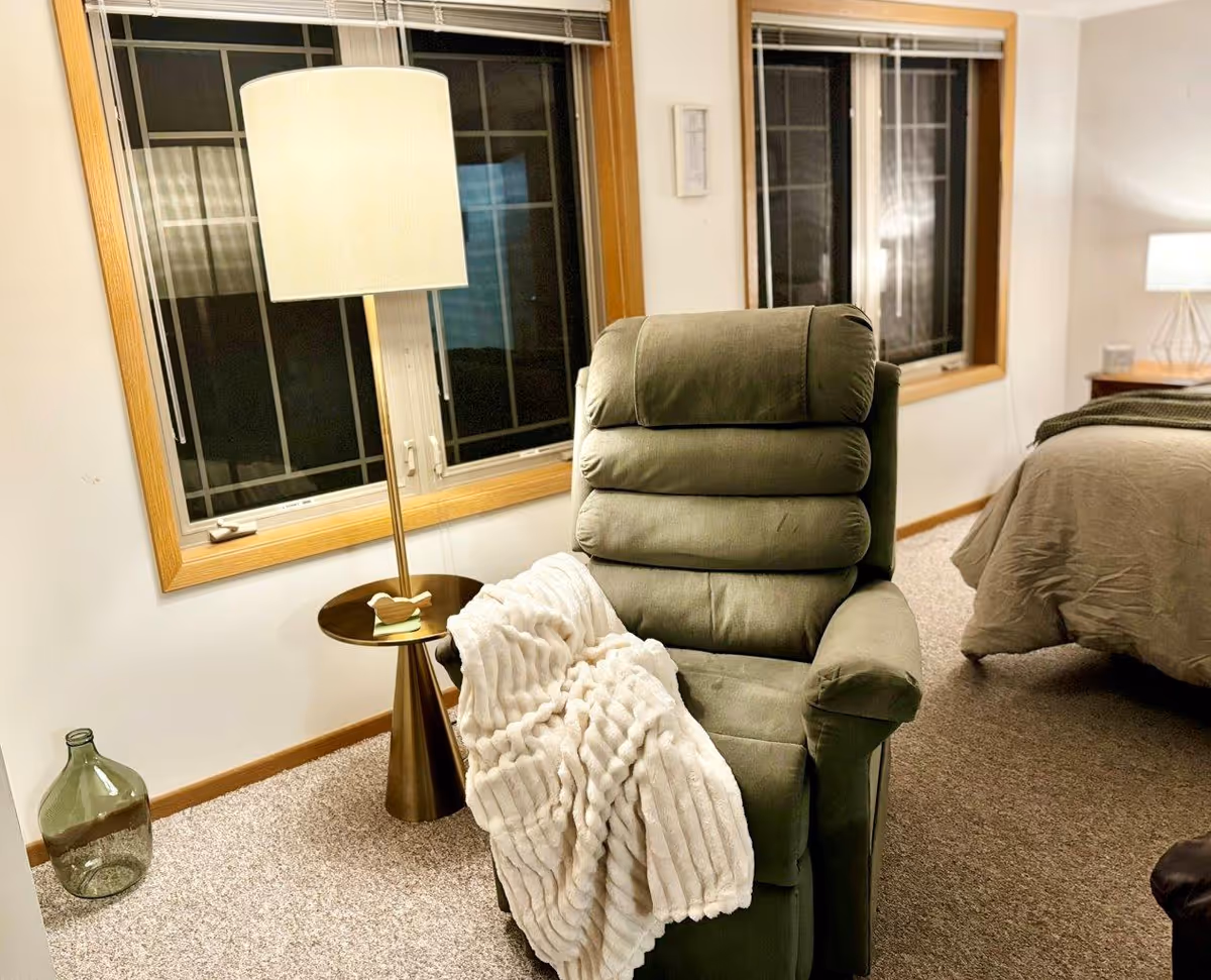 A cozy interior room featuring a green cushioned recliner chair with a white textured throw blanket draped over one arm. Next to the chair is a modern floor lamp with a round side table attached, holding a small decorative bird. Behind the chair are two large windows with wooden frames. Part of a bed with brown bedding and a nightstand with a lamp are visible in the background. The room has beige carpet flooring.