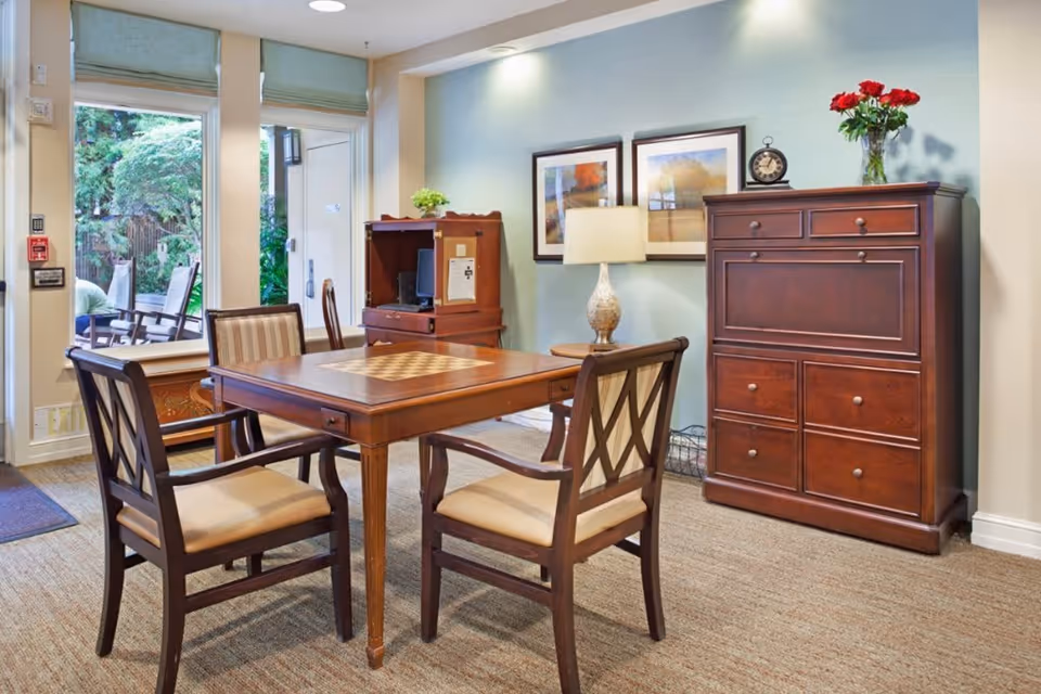 A cozy interior room with a wooden table featuring a built-in chessboard, surrounded by four cushioned wooden chairs. Against the wall is a wooden cabinet with drawers and a vase of red flowers on top. Next to it is a small desk with a computer and a lamp on a side table. Two framed pictures hang on the light blue wall. Large windows and a glass door reveal an outdoor patio with chairs and greenery.