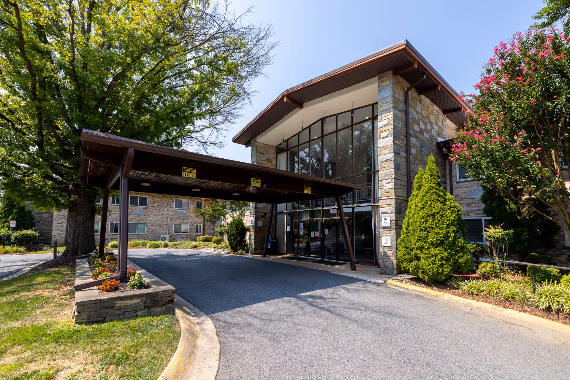 Exterior view of Autumn Lake Healthcare at Chevy Chase showing the main entrance with a covered drop-off area, large glass windows, stone facade, and surrounding greenery including trees and shrubs.