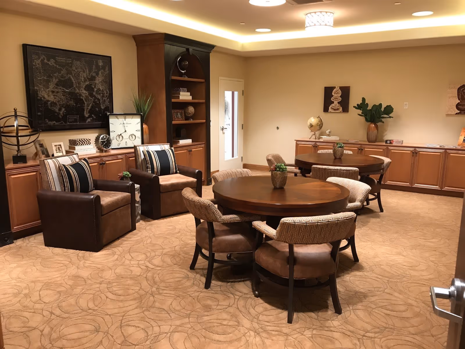 A cozy interior room with two round wooden tables each surrounded by four cushioned chairs. Two brown leather armchairs with striped pillows are positioned near a wooden cabinet with decorative items including a globe, clock, and framed pictures. The room has beige walls, carpeted floor with circular patterns, recessed ceiling lights, and decorative plants and artwork on the walls.