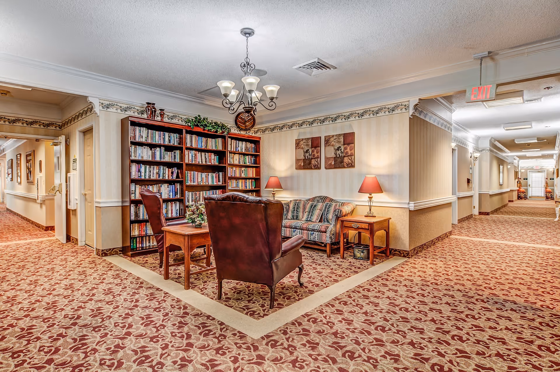 A cozy seating area in a senior living facility hallway featuring a bookshelf filled with books, a striped sofa, two leather armchairs, a wooden coffee table, and two side tables with red lamps. The area is carpeted with a patterned design, and the walls are decorated with wallpaper and framed artwork. The hallway extends on both sides with doors and lighting fixtures visible.