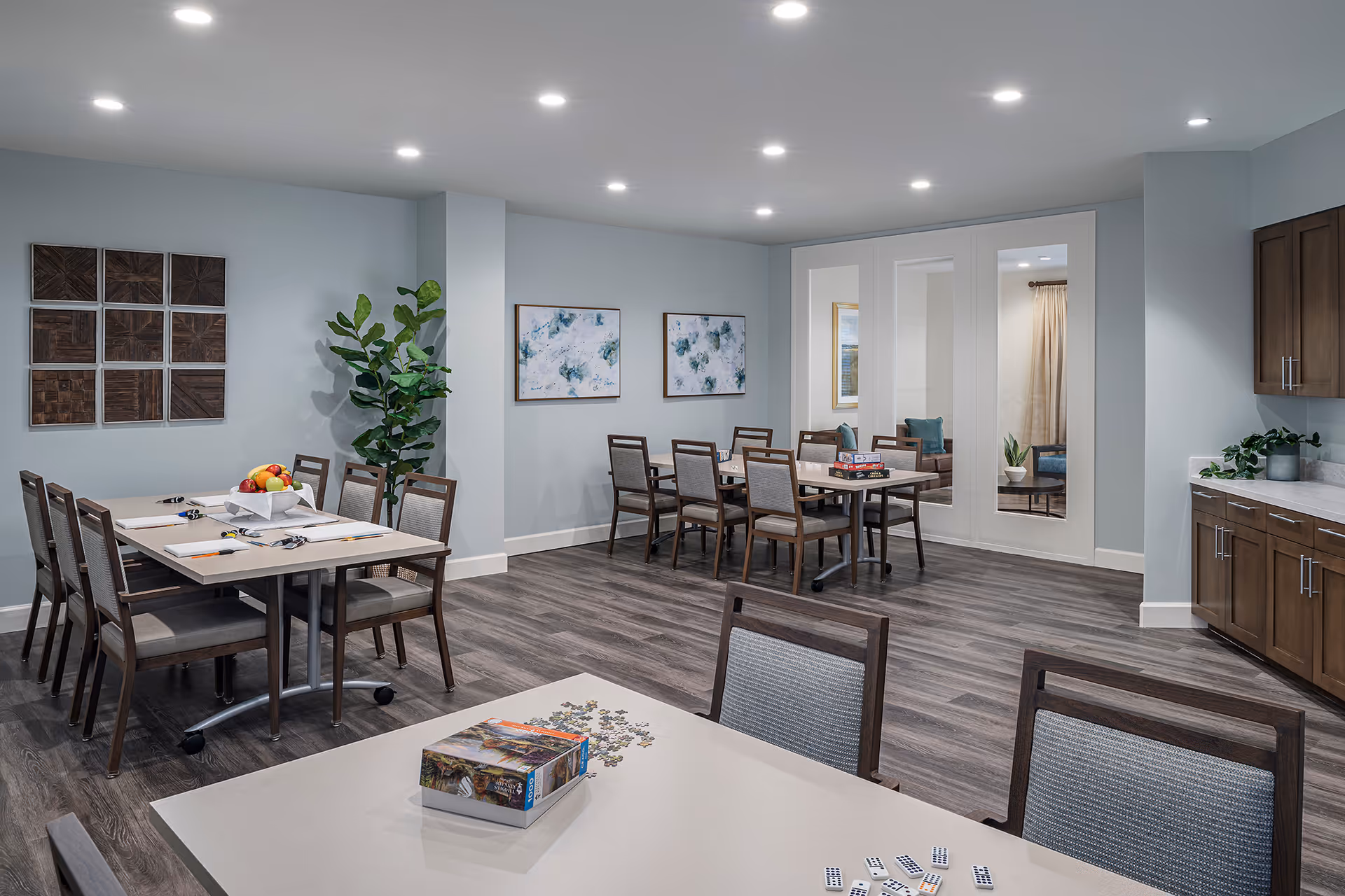 Bright communal dining/activity room with multiple tables and chairs, cabinets, wall art, and a potted plant.