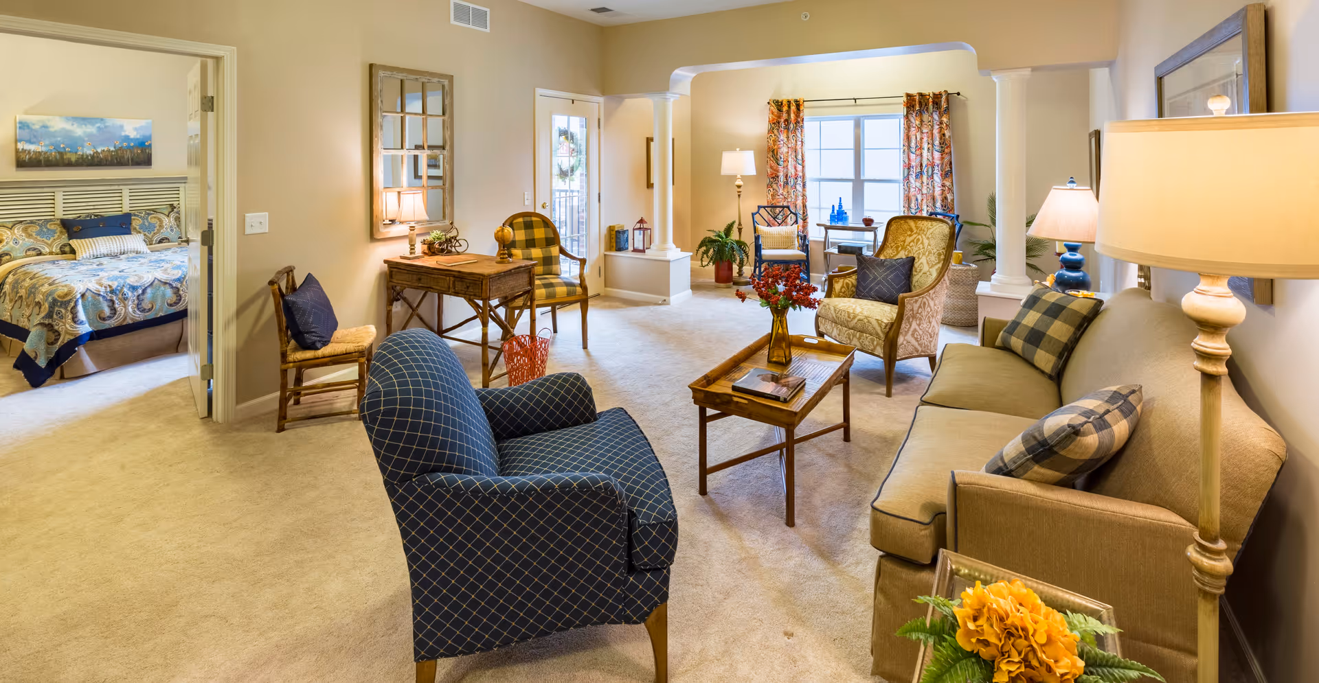A cozy living room area with a beige sofa, blue patterned armchair, wooden coffee table with a vase of flowers, and additional seating including a yellow armchair and a wooden chair. The room has beige carpet and walls, with a window featuring floral curtains letting in natural light. To the left, an open doorway reveals a bedroom with a bed covered in a patterned bedspread and a landscape painting above the headboard.