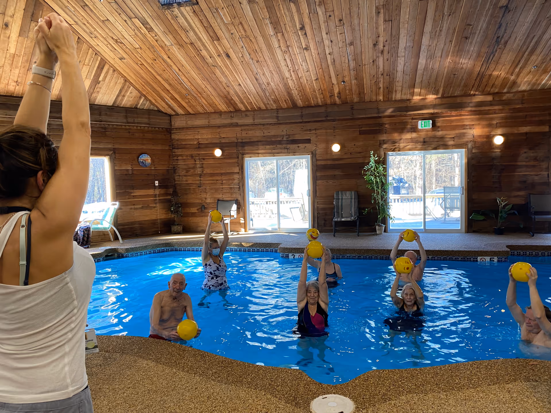 A group of elderly people participating in a water exercise class in an indoor pool with wooden walls and ceiling. An instructor stands at the poolside with arms raised, leading the class. The participants are holding yellow balls above their heads while standing in the water. Large windows and glass doors let in natural light, and there are chairs and plants around the pool area.