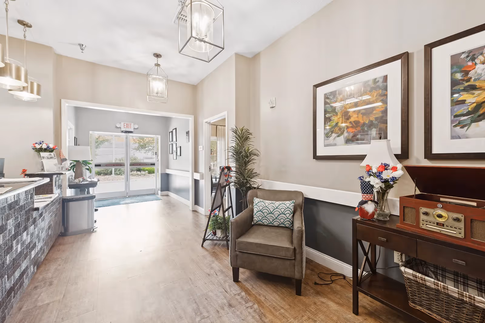 Bright reception lobby with a front desk, armchair and side table with lamp and flowers, and glass entrance doors in the background.