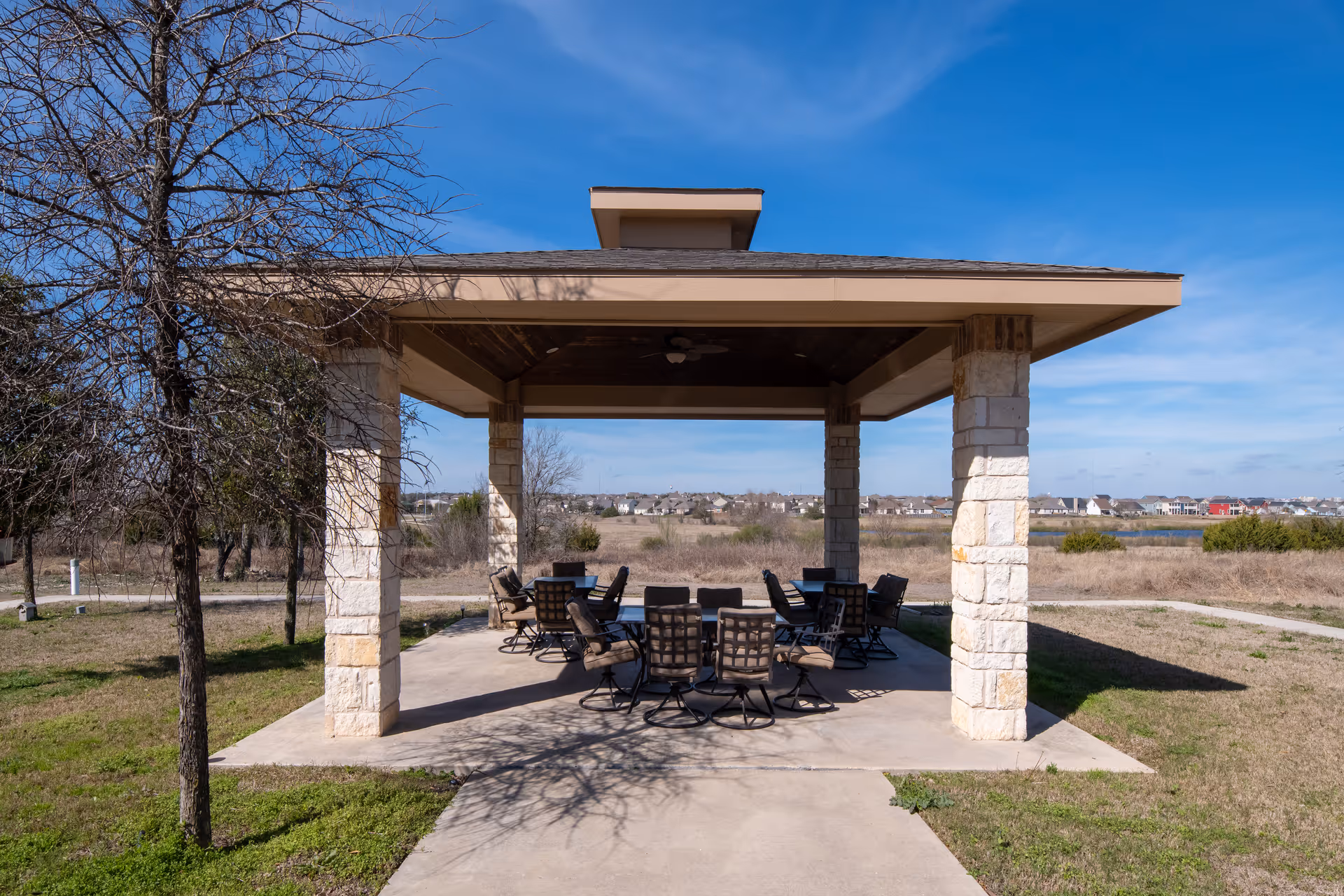 Outdoor covered pavilion with stone pillars and a roof, furnished with multiple cushioned chairs arranged around tables, situated in a grassy area with a clear blue sky and distant houses in the background.