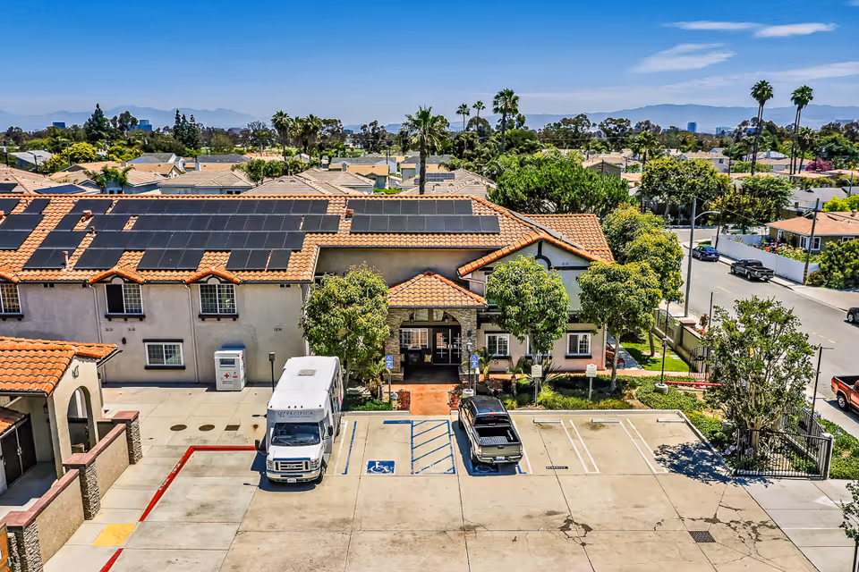 Aerial view of a senior living facility with a terracotta tiled roof equipped with solar panels. The building has a central entrance with a small covered porch, surrounded by trees and greenery. In front of the entrance, there is a parking lot with a white shuttle van and a black pickup truck parked in designated spaces, including a handicapped parking spot. The surrounding area includes residential houses and palm trees under a clear blue sky.