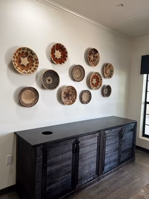 A black wooden cabinet with four doors sits against a white wall decorated with a collection of eleven woven decorative baskets arranged in a pattern. To the right, there is a window with black trim allowing natural light into the room.