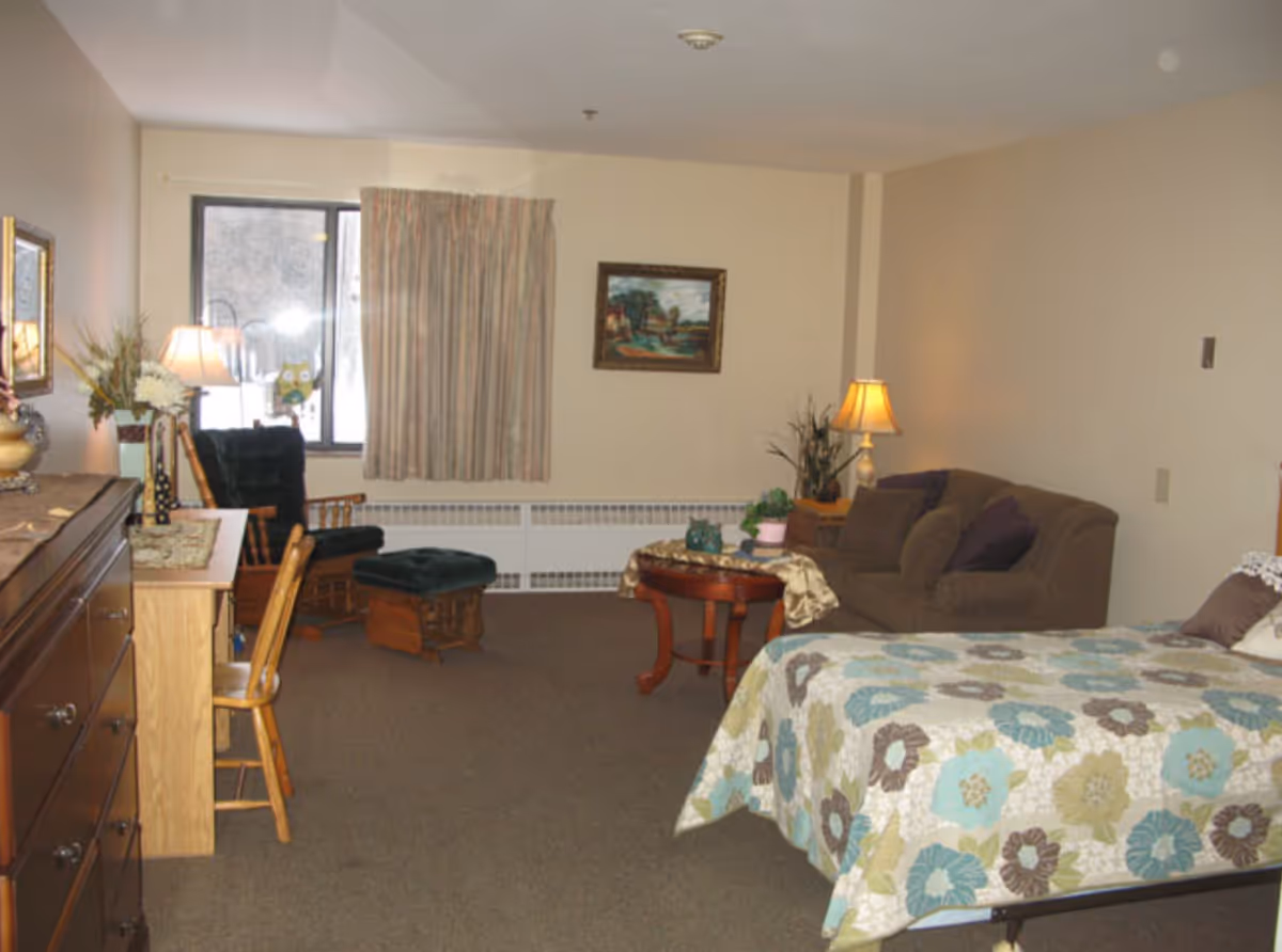 A senior living facility room with a bed covered in a floral quilt, a brown couch with pillows, a wooden coffee table with plants and decorative items, a wooden rocking chair with a matching footrest, a wooden dresser, a desk with a chair, two table lamps, a window with curtains, and a framed painting on the wall.