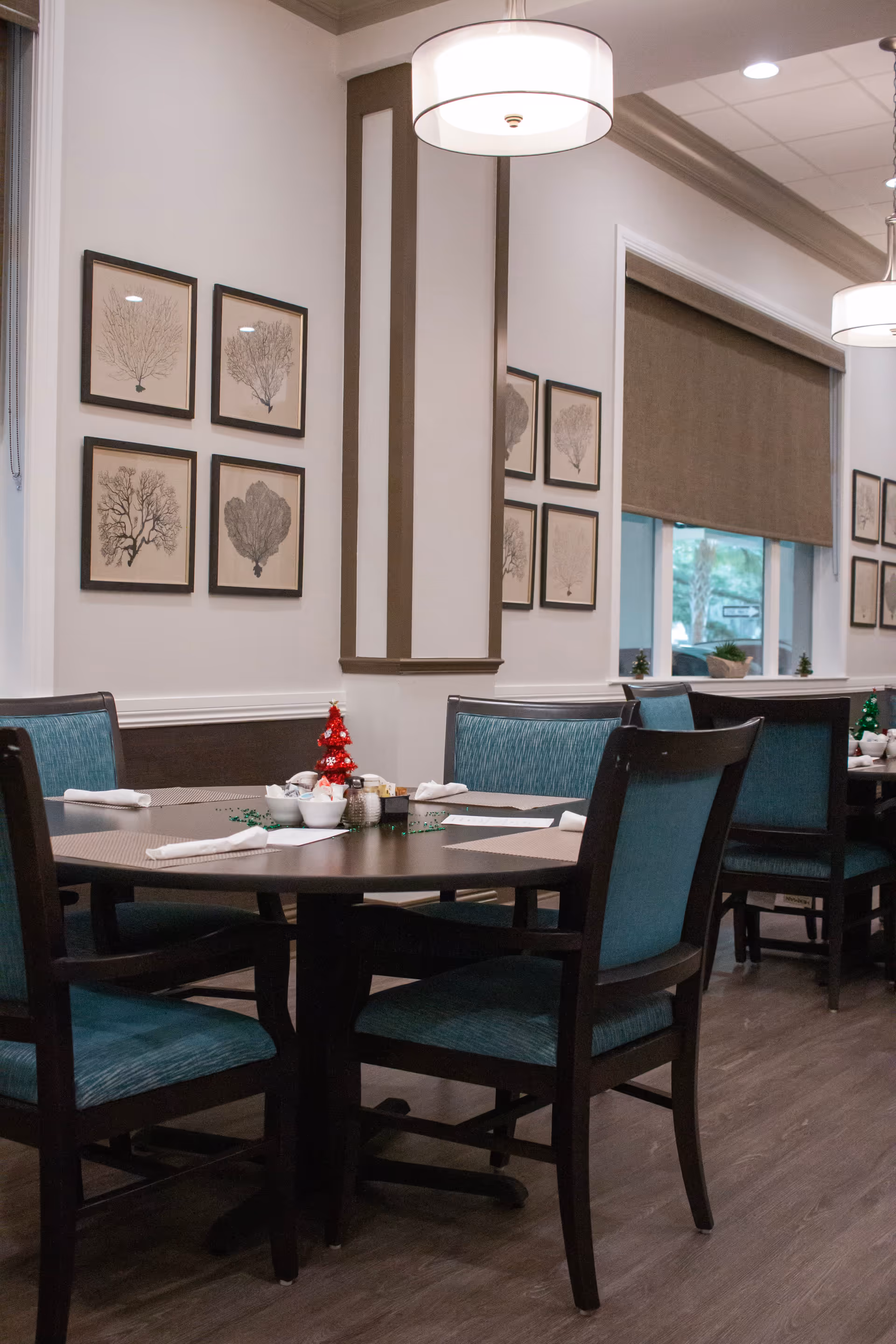 Interior dining area with round tables and teal cushioned chairs. Tables are set with placemats, napkins, and small bowls. The walls are decorated with framed botanical prints, and there are large windows with brown blinds. Modern pendant lights hang from the ceiling.