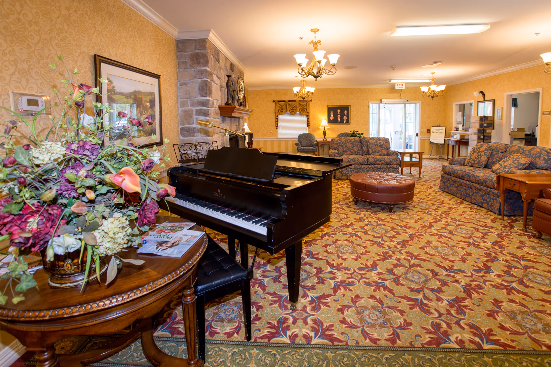 A spacious living room area with patterned carpet, floral upholstered sofas, a round leather ottoman, and a black grand piano. There is a wooden table with a large floral arrangement and magazines on it. The room features a stone fireplace, framed artwork on the walls, and multiple chandeliers providing warm lighting. The back of the room has a window with curtains and a glass door leading outside.