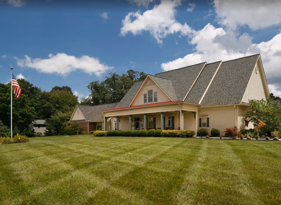 A large, light-colored building with a steep roof and a covered front porch, surrounded by a well-maintained lawn and bushes. An American flag on a flagpole stands on the left side of the image, with trees and a partly cloudy blue sky in the background.