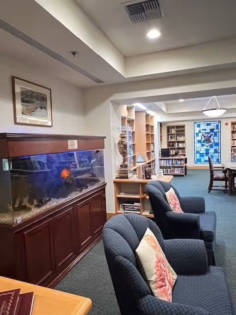 Interior view of a senior living community common area featuring a large fish tank on a wooden cabinet, two blue upholstered armchairs with floral pillows, bookshelves filled with books, a stained glass window, and a table with chairs in the background.