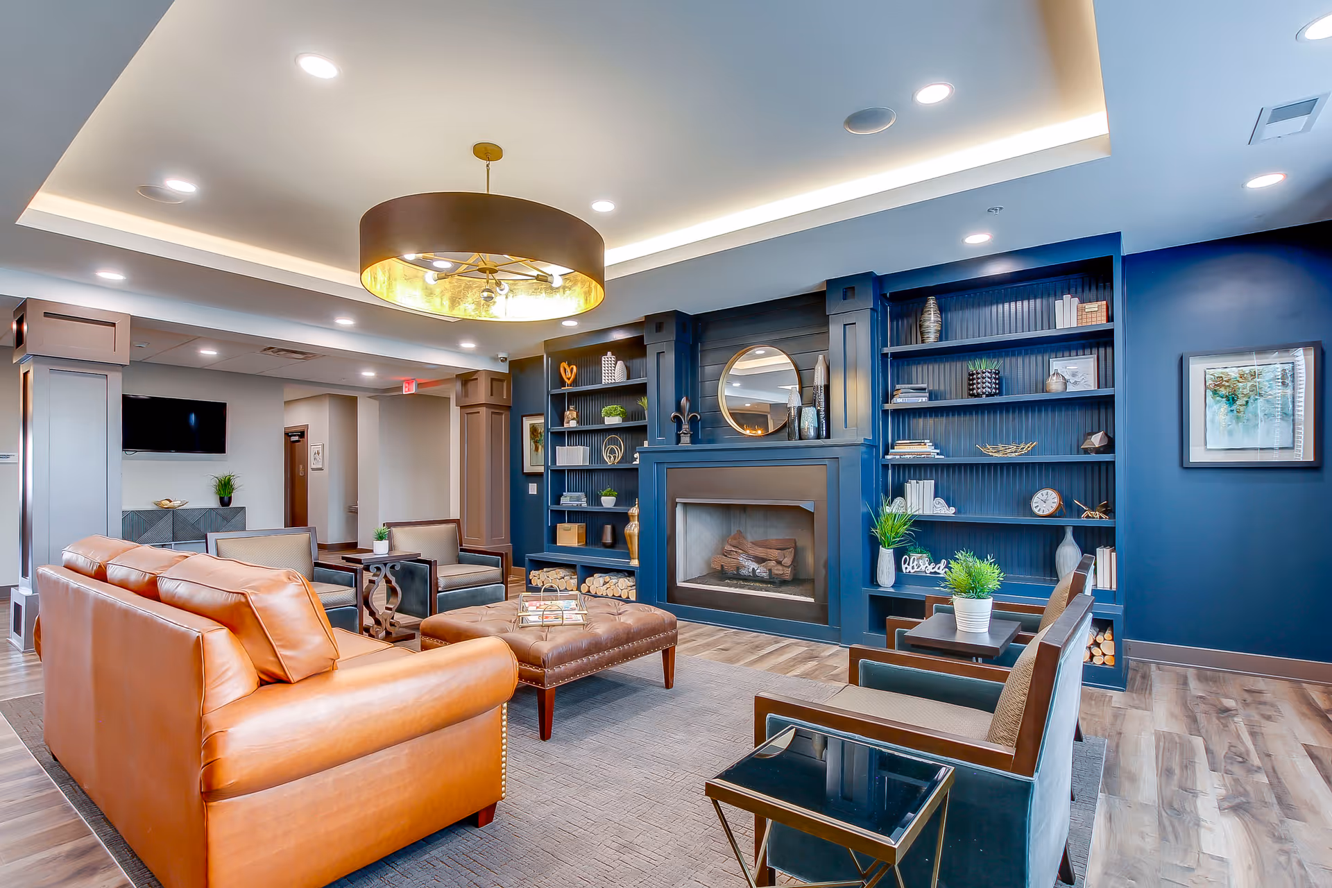 Bright communal lounge with leather sofas, armchairs, a central ottoman, and a fireplace flanked by built-in blue shelves.