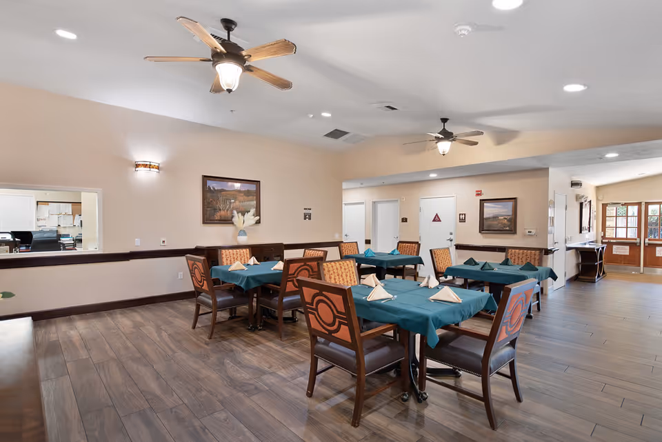 Dining room with multiple tables set with green tablecloths and folded napkins, ceiling fans, and wood-look flooring.