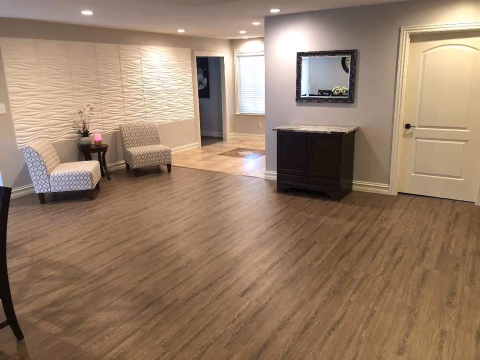 A spacious interior room with wood flooring, two patterned chairs with a small table between them holding a plant and a small lamp, a textured white wall panel, a dark wooden cabinet with a marble top beneath a framed mirror, and a closed white door. The room is well-lit with recessed ceiling lights.