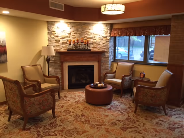 Cozy seating area with four upholstered armchairs arranged around a round ottoman in front of a stone fireplace and window.