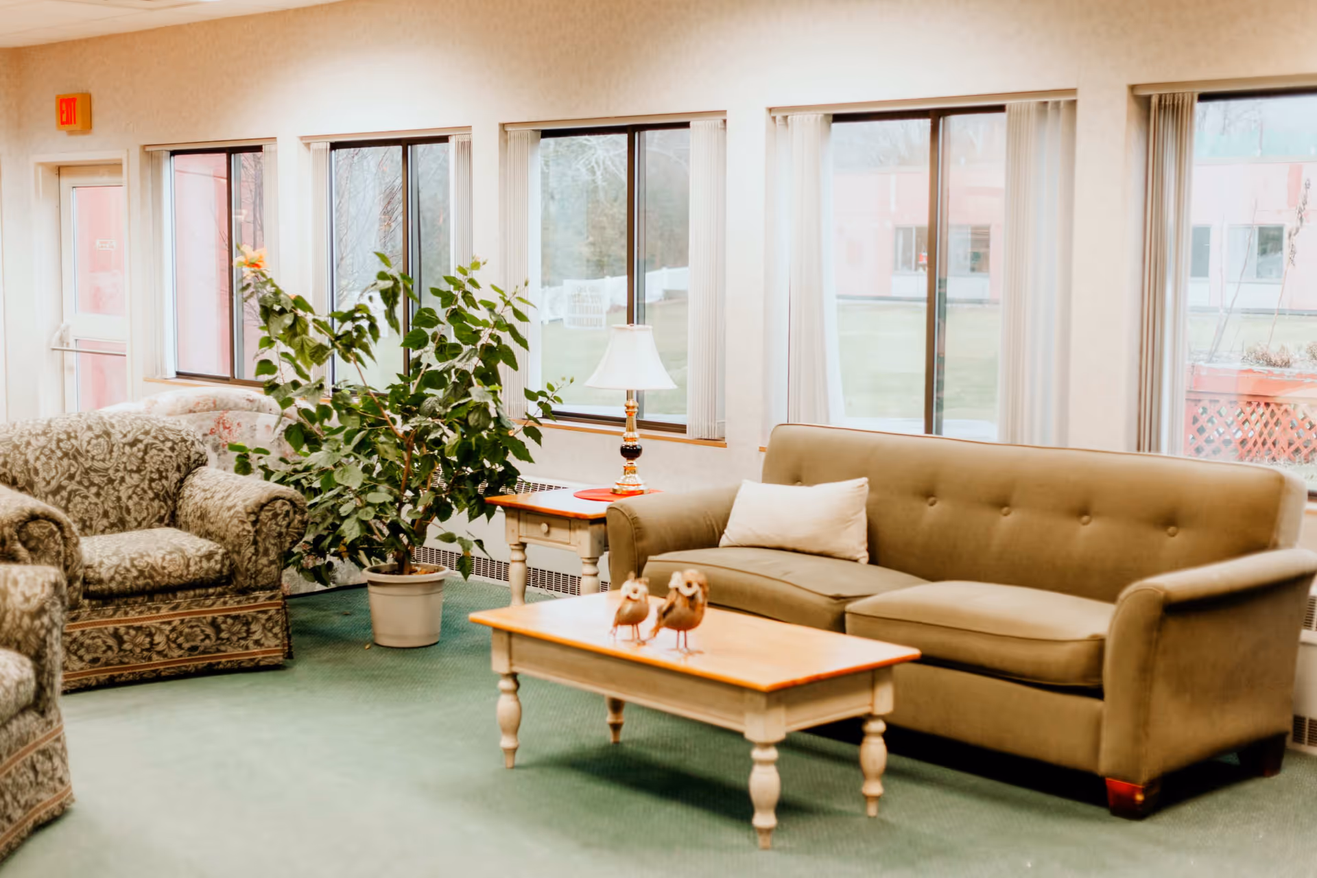 A cozy senior living community lounge area with a green upholstered sofa, a patterned armchair, a wooden coffee table with two bird figurines, a side table with a lamp, and a large potted plant. Large windows with vertical blinds let in natural light and show an outdoor view.