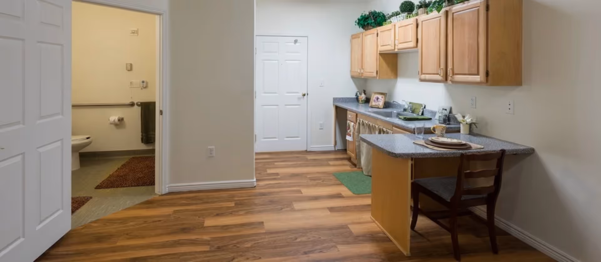 Interior view of a senior living facility kitchen area with wooden cabinets, a countertop with a place setting, and a chair. To the left, there is an open door leading to a bathroom with a toilet, grab bars, and a towel hanging on the wall. The floor is wood laminate in the kitchen and tile in the bathroom.