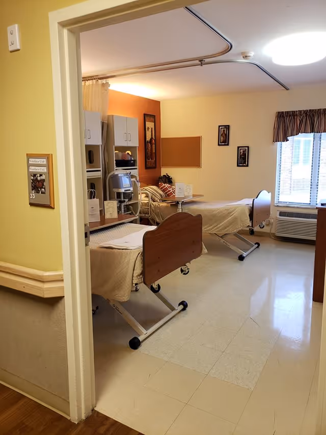 A room with two hospital-style beds with beige bedding, medical equipment, and cabinets along the wall. There is a window with blinds and a valance, framed pictures on the walls, and a bulletin board. The floor is tiled and the walls are painted in light yellow and orange tones.
