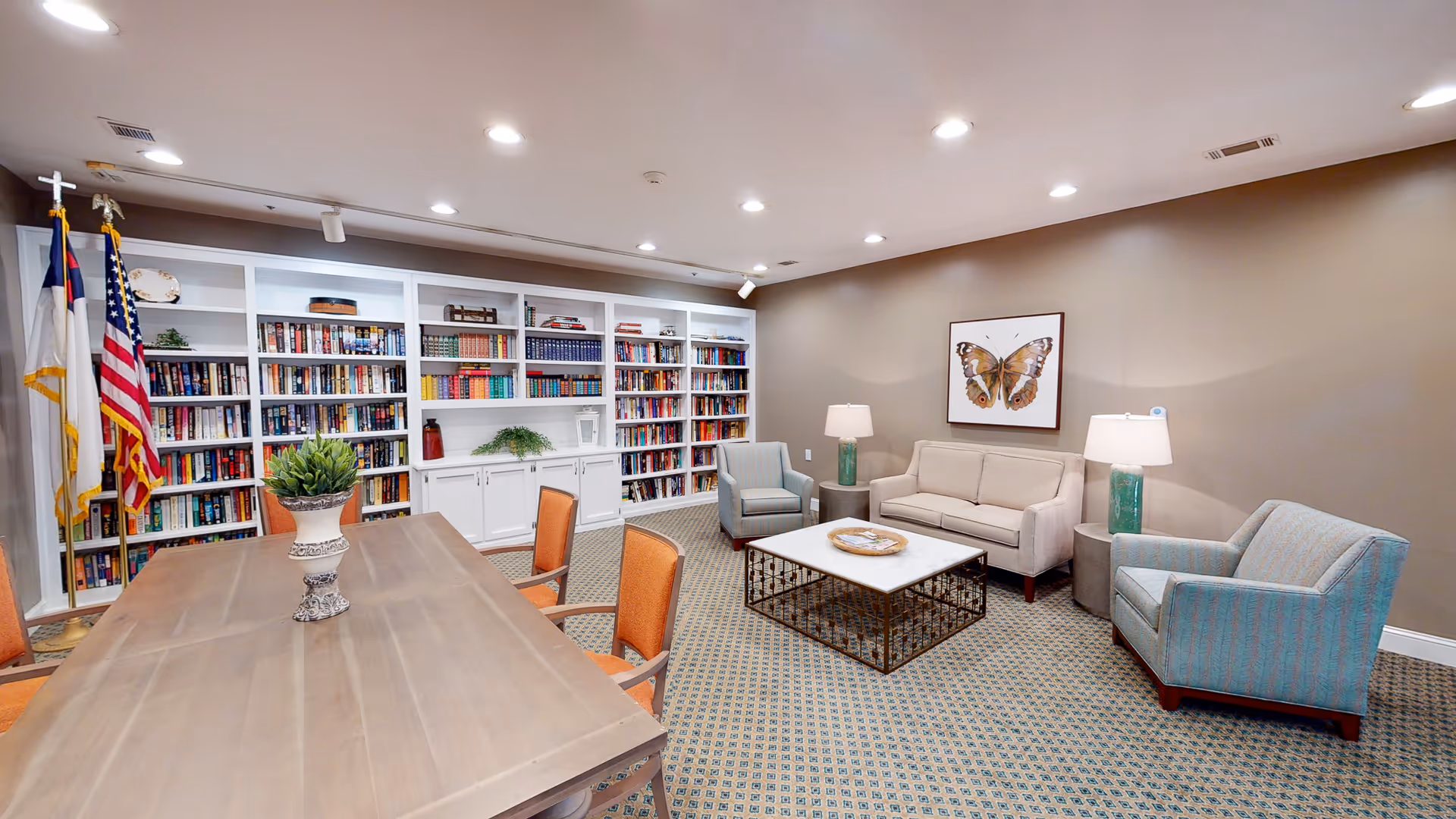 A cozy library and sitting area in a senior living facility featuring a large wooden table with orange chairs, a white bookshelf filled with books, two flags in the corner, a beige sofa, two blue armchairs, a white marble coffee table, two side tables with lamps, and a framed butterfly artwork on the wall.