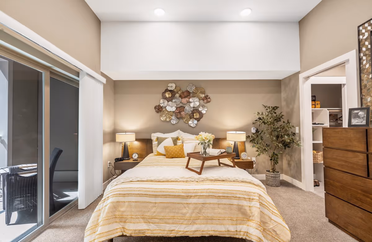 A cozy bedroom with a bed covered in a yellow and white striped blanket, two bedside tables with lamps, a decorative metal wall art above the bed, a wooden dresser, a potted plant, and a sliding glass door leading to an outdoor patio with chairs.