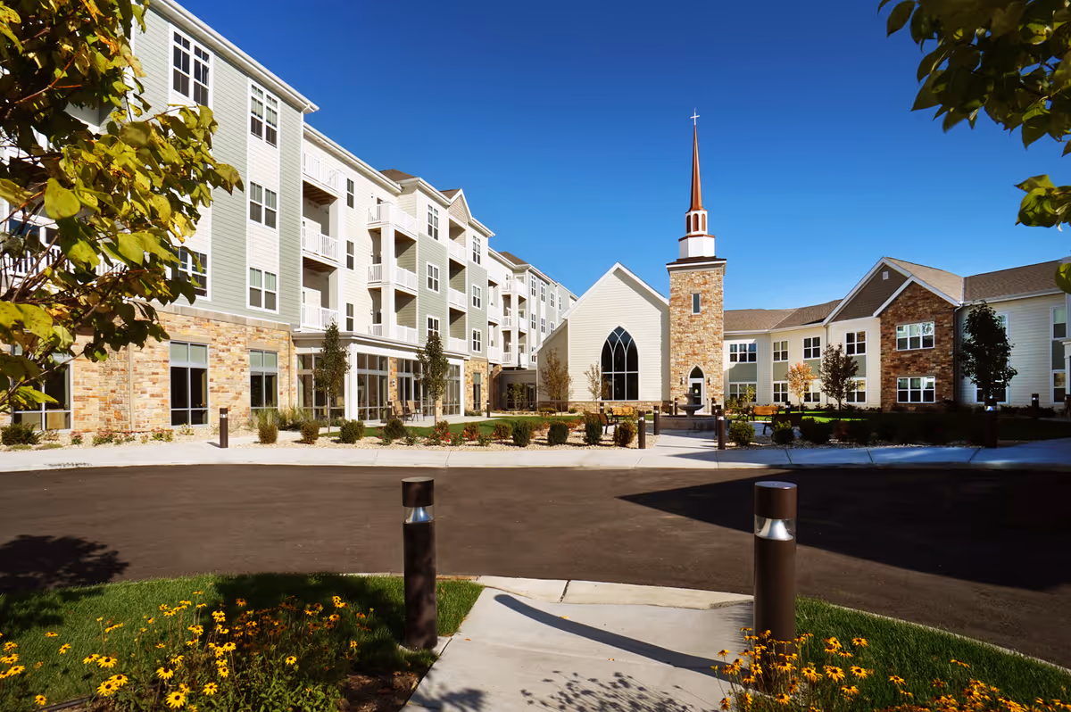 Exterior view of a senior living facility named All Saints Neighborhood featuring a multi-story building with balconies, a church-like structure with a tall steeple and cross, landscaped gardens with flowers and trees, and a clear blue sky.