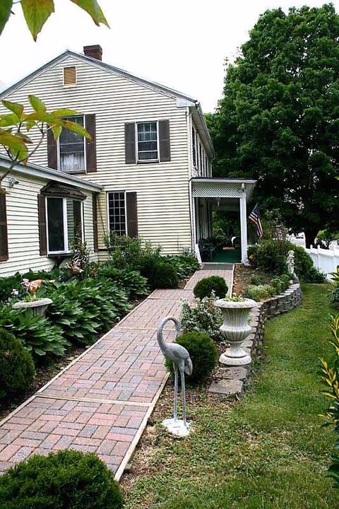 Two-story beige house with a brick walkway, garden beds, and a small front porch.
