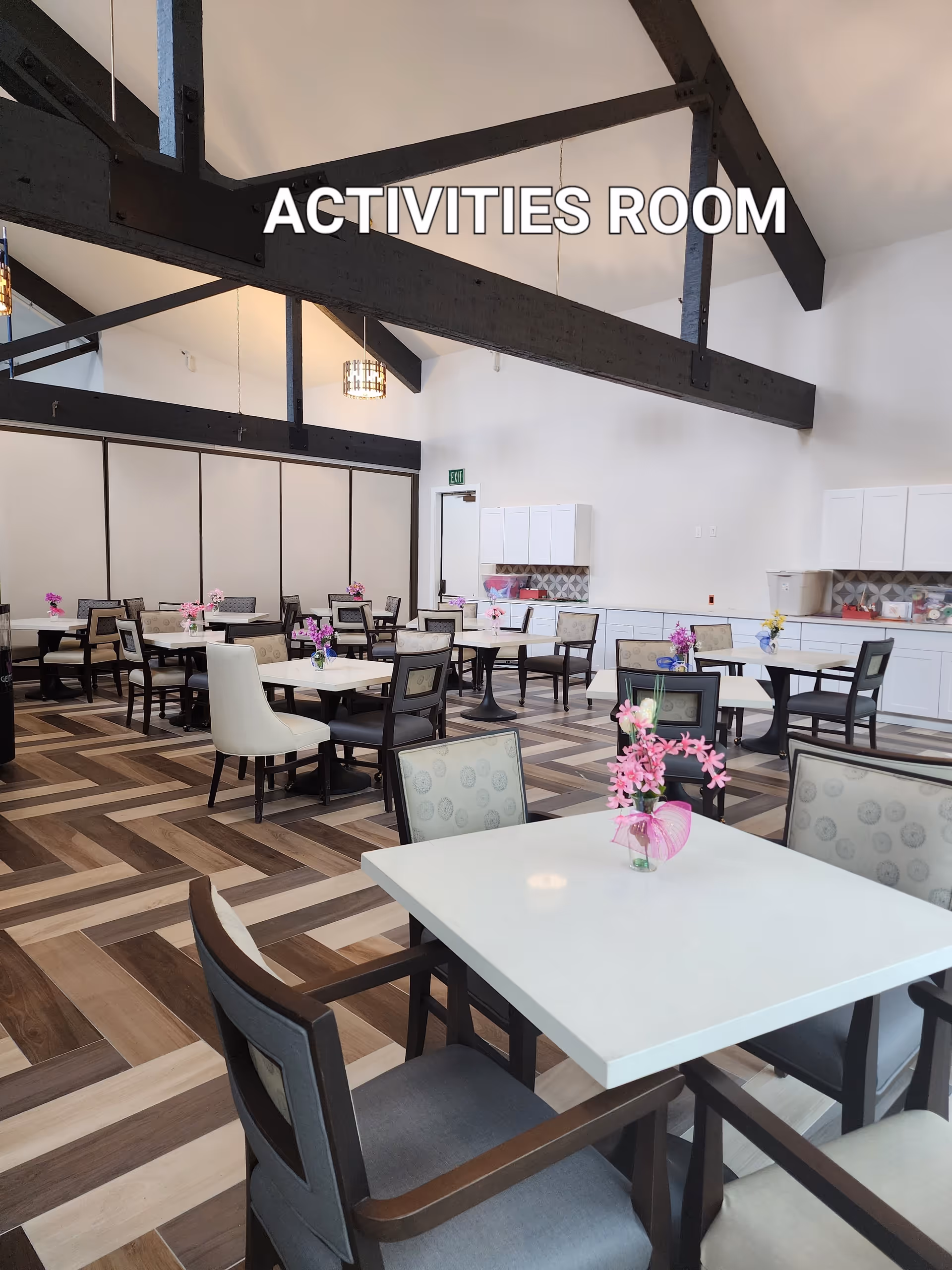 Spacious activities room with multiple tables and chairs arranged neatly. Each table has a small vase with colorful flowers. The room features high ceilings with exposed dark wooden beams and modern pendant lights. The floor has a patterned wood design, and there are white cabinets along one wall.