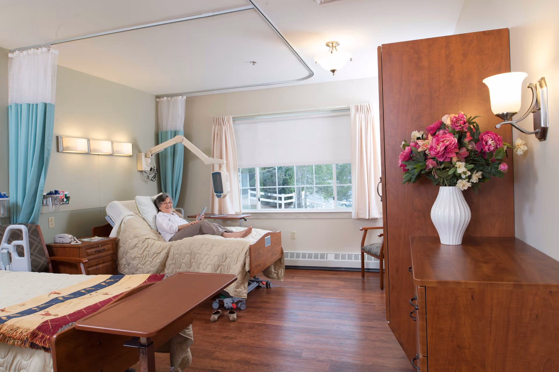 A senior woman reclining on a hospital-style bed in a well-lit room with wooden flooring. The room has a large window with curtains, a wooden cabinet with a vase of pink and white flowers, a bedside table with a telephone, and a chair near the window. Medical equipment is visible near the bed.