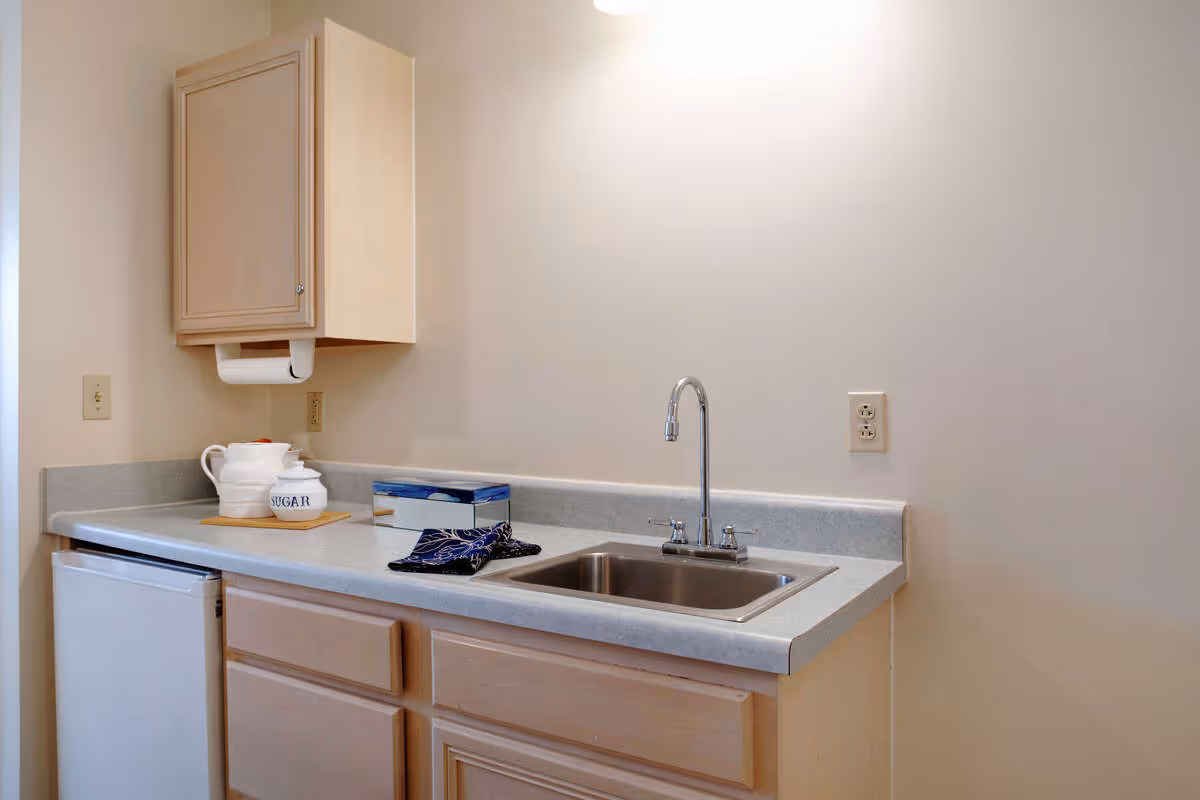 A small kitchenette area with a light-colored countertop, a stainless steel sink with a faucet, light wood cabinets above and below the counter, a mini refrigerator, a white teapot and sugar container on a wooden tray, a box of tissues, and a folded dark cloth on the counter.