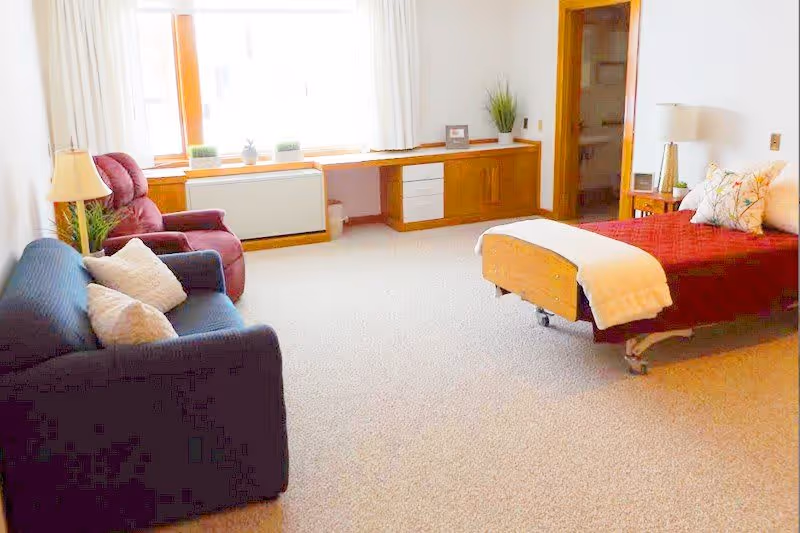 A bright and spacious room with a hospital-style bed covered with a red blanket and white throw, a blue couch with pillows, a red recliner chair, a wooden desk with drawers under a large window, and a lamp on a side table. The room has beige carpet and white walls.