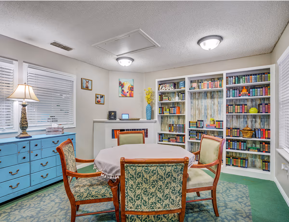 A cozy room with a round table covered by a light-colored tablecloth surrounded by four wooden chairs with patterned upholstery. Behind the table, there is a white bookshelf filled with colorful books and decorative items. To the left, a blue dresser with multiple drawers holds a lamp and a tissue box. The walls are decorated with three small paintings of animals, and the room is lit by ceiling lights and natural light from windows with white blinds.