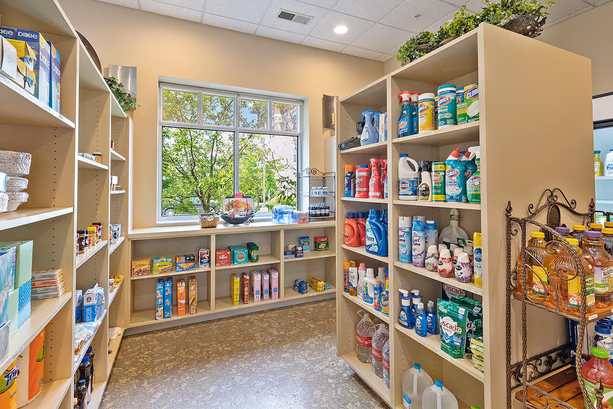 A well-lit room with shelves stocked with various household and cleaning products, snacks, and bottled beverages. A large window lets in natural light and shows green trees outside. The floor is a polished concrete surface.