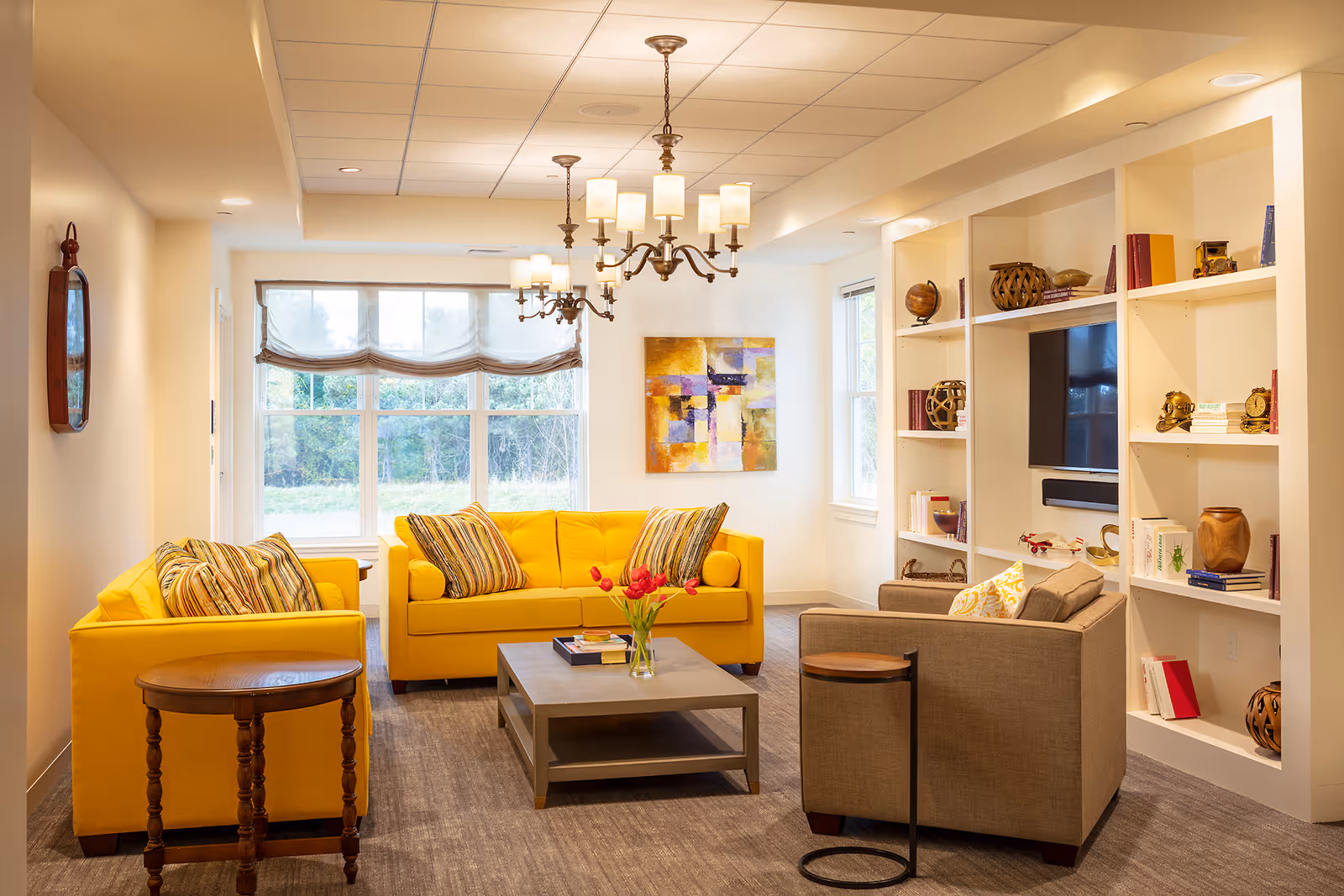 A bright and cozy living room with two yellow sofas and one beige armchair arranged around a gray coffee table with a vase of red tulips. The room features large windows with sheer curtains, built-in white shelves filled with books and decorative items, a wall-mounted flat-screen TV, and two elegant chandeliers hanging from the ceiling. A colorful abstract painting is displayed on the wall.
